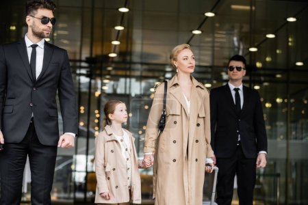 two bodyguards walking next to successful woman and preteen kid, entering hotel, private security, blonde mother and daughter in trench coats, safety and protection, family travel, rich life 