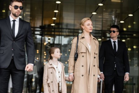 Photo for Bodyguards walking next to blonde successful woman and preteen kid, entering hotel, private security, mother and daughter in trench coats, safety and protection, family travel, rich life - Royalty Free Image