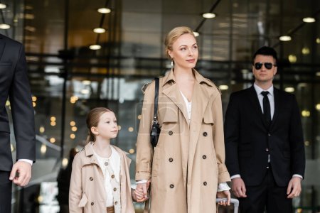 Photo for Bodyguards walking next to blonde woman and preteen kid, entering hotel, private security, successful mother holding hands with daughter and wearing trench coats, safety and protection - Royalty Free Image