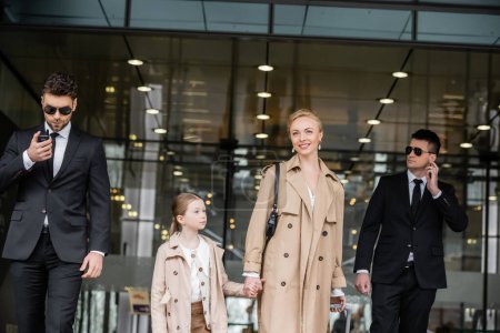 bodyguards walking next to cheerful woman and preteen kid, entering hotel, private security, mother holding hands with daughter and wearing trench coats, safety and protection 