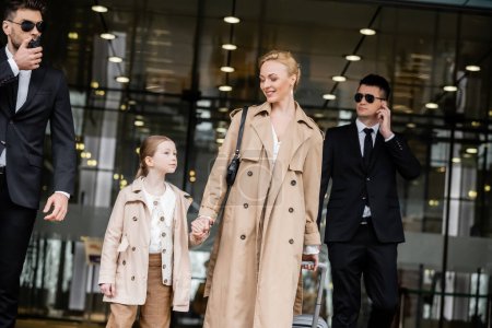 bodyguards communicating with each other next to happy woman and preteen kid, entering hotel, private security, mother holding hands with daughter and wearing trench coats, safety and protection 