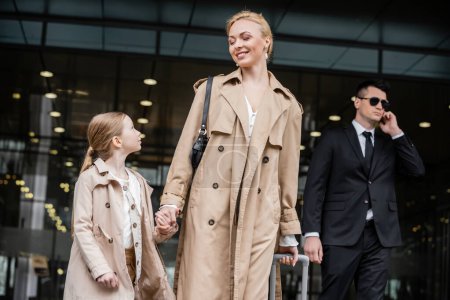 personal security, lifestyle, blonde mother with preteen girl holding hands near hotel, successful woman and child, bodyguard in suit standing on blurred background, rich life 