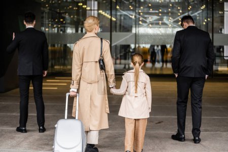back view of two bodyguards walking next to blonde woman and child, entering hotel, private security, mother holding hands with preteen daughter, life safety and protection 