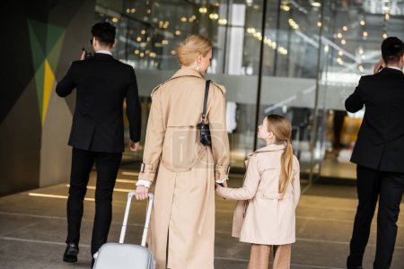bodyguards walking next to blonde woman and preteen kid, entering hotel, private security, mother holding hands with daughter, people wearing trench coats, safety and protection, back view 