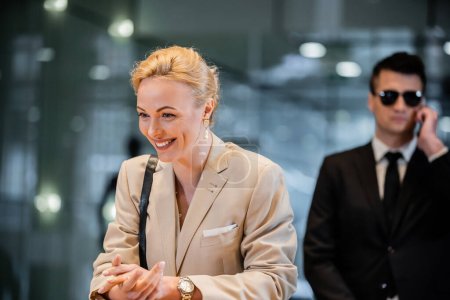 happy blonde woman in formal wear standing at reception desk, personal security service concept, bodyguard in suit on blurred background, hotel industry, luxury travel, formal wear 
