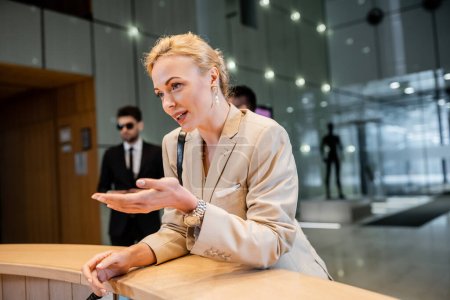 blonde woman in suit gesturing while talking at reception desk, personal security service concept, bodyguards in suits standing on blurred background, hotel industry, safety during travel 