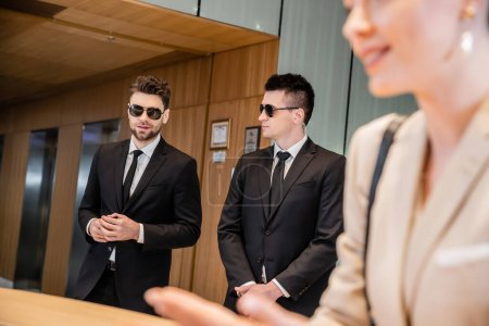 Photo for Handsome bodyguards protecting successful client in hotel, woman on blurred foreground, security service, personal safety, men in suits and sunglasses, hotel reception - Royalty Free Image