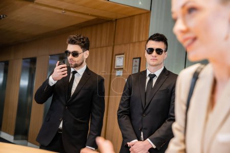 handsome bodyguards protecting beautiful and successful client in hotel, blonde woman on blurred foreground, security service, personal safety, man using walkie talkie 