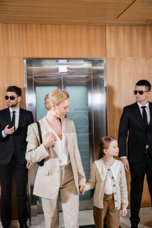mother and child holding hands while standing near hotel elevators and bodyguards in suits, personal protection, successful woman and preteen daughter, family travel, security service 