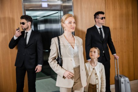 successful mother and child holding hands while standing near hotel elevator and bodyguards in suits, personal protection, woman and preteen daughter, family travel, security service 