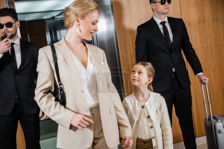 joyful mother and child holding hands while standing near hotel elevator and bodyguards in suits, personal protection, successful woman and preteen daughter, family travel, security service 