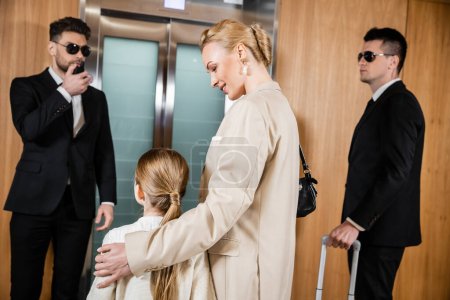 blonde mother hugging child while standing near hotel elevator and bodyguards in suits, personal safety and protection, successful woman and preteen daughter, family travel, security service 