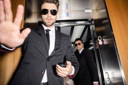 private security concept, handsome bodyguard in sunglasses and suit showing stop, no gesture to camera, protecting private space, hotel lobby, standing next to work partner in elevator 