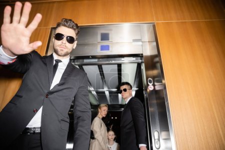 private security concept, handsome bodyguard in sunglasses and suit showing stop, no gesture to camera, protecting private space of famous clients in elevator of luxury hotel, mother and daughter 
