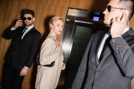 personal security and protection concept, successful blonde woman with handbag standing near elevator next to bodyguards in suits and sunglasses, luxury hotel, female guest 