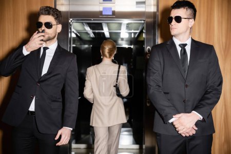 persönliches Sicherheits- und Schutzkonzept, blonde und erfolgreiche Frau mit Handtasche im Fahrstuhl neben Leibwächtern in Anzug und Sonnenbrille, Luxusleben, Hotelgast 