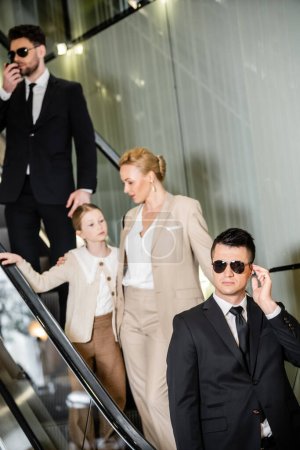 Photo for Personal security concept, handsome bodyguards communicating while protecting safety of female clients, rich lifestyle, blonde and successful woman and preteen girl standing on escalator of hotel - Royalty Free Image