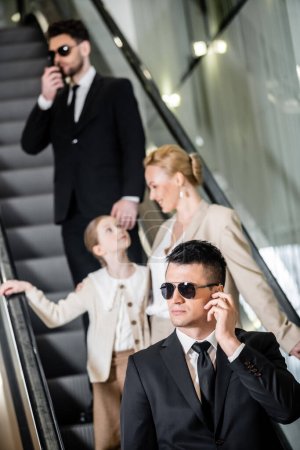 Photo for Personal security concept, handsome bodyguards communicating while protecting safety of female clients, rich lifestyle, successful woman and preteen child standing on escalator of hotel - Royalty Free Image