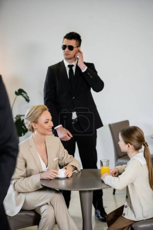 privates Sicherheitskonzept, erfolgreiche glückliche Mutter und Tochter verbringen Zeit miteinander im Café, trinken Kaffee und Orangensaft, zwei Leibwächter schützen die Sicherheit weiblicher Kunden 