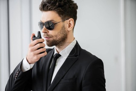 Photo for Surveillance, handsome bodyguard communicating through walkie talkie, man in sunglasses and black suit with tie, hotel safety, security management, uniformed guard on duty, professional headshots - Royalty Free Image