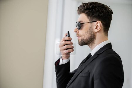 Photo for Surveillance, bodyguard communicating through walkie talkie, man in sunglasses and black suit with tie, hotel safety, security management, uniformed guard on duty, professional headshots, side view - Royalty Free Image