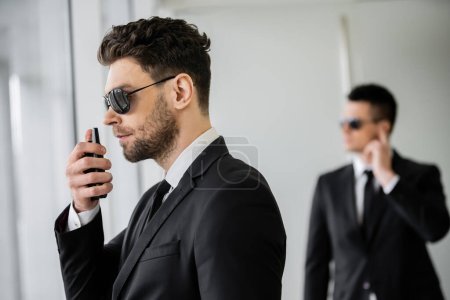 surveillance, bodyguard communicating through walkie talkie, man in sunglasses and black suit with tie, hotel safety, security management, uniformed guard on duty, work partner on background 
