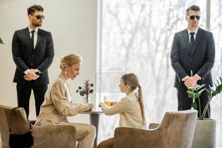 Photo for Private security concept, bodyguards standing near female clients, preteen girl and blonde mother spending time together in cafe, personal safety and protection, professional in suits - Royalty Free Image