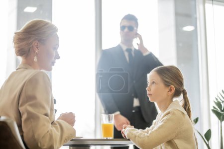 preteen girl talking to blonde mother near bodyguard on blurred background, security service, private safety concept, glass of orange juice, personal safety 