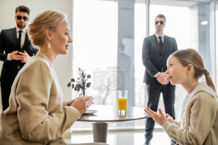 Photo for Security service, private safety concept, preteen girl talking with blonde mother over drinks in cafe, bodyguards standing on blurred background, personal safety, spending time together - Royalty Free Image