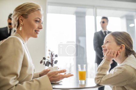 security service, private safety concept, cheerful blonde woman with cup of coffee talking with preteen daughter in cafe, bodyguards on blurred background, beverages and communication 