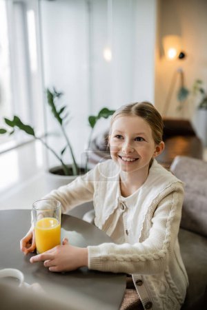 happy preteen girl holding glass of orange juice while sitting in cafe, positivity and joy, round table, smart casual attire, blurred plant on background 