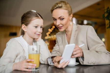 Mutter und Tochter verbringen Zeit miteinander, blonde Frau hält Smartphone in der Nähe der Tochter, berufstätige Eltern und Kind, moderne Elternschaft, familiäre Bindung, ausgewogener Lebensstil 