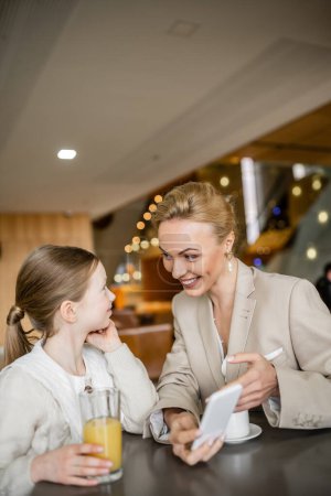 Photo for Happy mother and daughter spending quality time together, blonde woman holding smartphone near daughter, digital age, working parent and kid, modern parenting, family bonding - Royalty Free Image