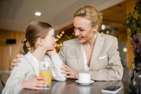 glückliche Mutter, die Zeit mit ihrer Tochter verbringt, blonde Frau, die ein junges Mädchen in der Nähe von Getränken und Smartphone umarmt, berufstätige Eltern und Kind, moderne Elternschaft, familiäre Bindung 