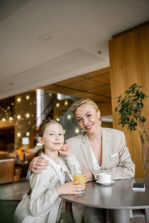 Photo for Mother and daughter spending quality time together, blonde woman hugging preteen girl and looking at camera near beverages and smartphone, working parent and kid, modern parenting, family bonding - Royalty Free Image