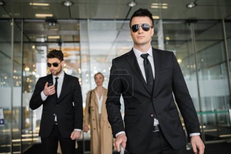 private security service, personal safety concept, handsome bodyguards in formal wear and sunglasses protecting safety of client on blurred background, walking in hotel 