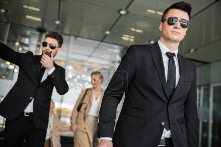 bodyguards protecting privacy of female clients, mother and daughter walking out of hotel on blurred background, personal safety of rich family, men in sunglasses and suit on duty 