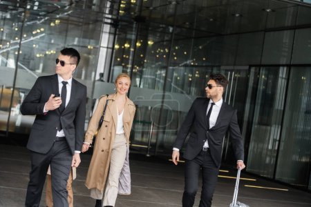 handsome bodyguards protecting privacy of female clients, happy woman and daughter walking out of hotel, personal safety of rich family, men in sunglasses and suit with luggage  