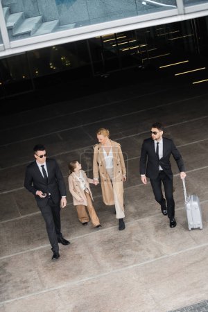 private security, top view of personal bodyguards protecting female clients, blonde woman and kid walking out of hotel, safety of rich family, men in formal wear and sunglasses on duty 