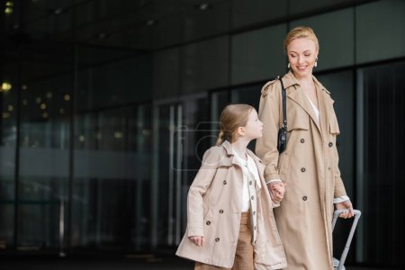 autumn fashion, mother daughter time, positive woman with luggage holding hand of preteen girl while walking out of hotel together, smart casual, beige trench coats, outerwear, modern parenting 
