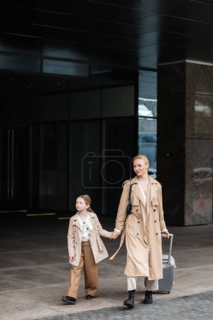autumn fashion, mother daughter time, happy woman with luggage holding hand of preteen kid while walking out of hotel together, smart casual, beige trench coats, outerwear, trendy look 