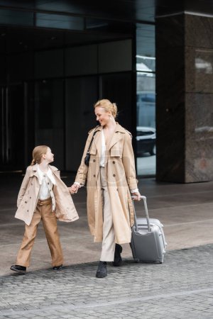 Photo for Trendy look, mother daughter time, happy woman with luggage holding hand of preteen girl while walking out of hotel together, smart casual, beige trench coats, outerwear, modern parenting - Royalty Free Image