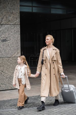 Photo for Outerwear, mother daughter time, happy woman with luggage holding hand of preteen girl while walking out of hotel together, smart casual, beige trench coats, autumn fashion, modern parenting - Royalty Free Image