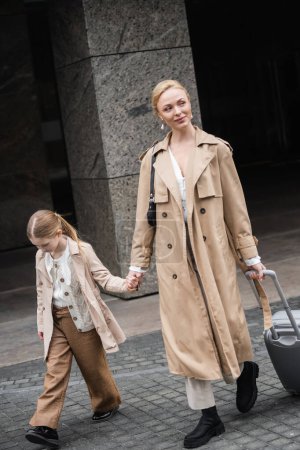 modern parenting, mother daughter time, happy woman with luggage holding hand of preteen girl while walking out of hotel together, smart casual, beige trench coats, outerwear, trendy look 