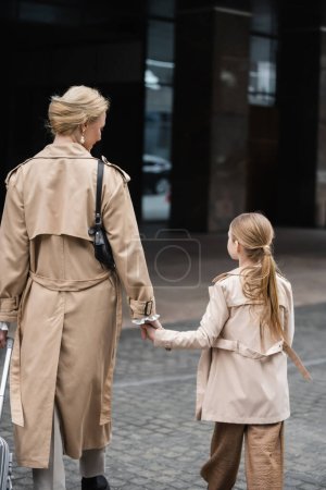 modern parenting, mother daughter time, back view of blonde woman with luggage holding hand of girl while walking out of hotel together, smart casual, beige trench coats, outerwear, trendy look 