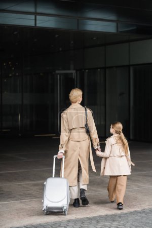 family travel, mother and child, back view of blonde woman with luggage holding hand of girl while walking into hotel together, smart casual, beige trench coats, outerwear, trendy look 