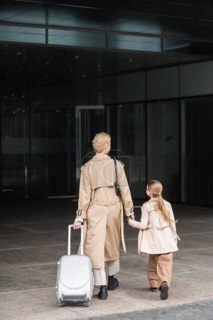 Familienreise, Mutter und Kind, Rückansicht einer blonden Frau mit Gepäck, die Hand des Mädchens hält, während sie gemeinsam ins Hotel geht, schicke lässige, beige Trenchcoats, Oberbekleidung, schicke Freizeitkleidung 