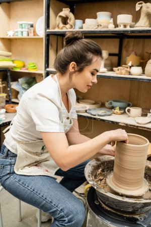 Téléchargez les photos : Vue latérale de l'artisan brune en tablier travaillant avec de l'argile sur roue de poterie en atelier en atelier - en image libre de droit