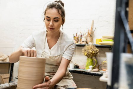 Brunette craftswoman in apron making clay vase and working on pottery wheel in ceramic workshop
