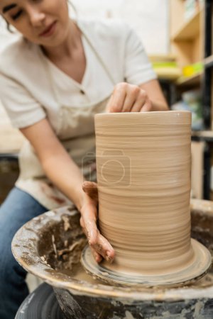 Vista recortada de alfarero hembra borrosa haciendo jarrón de arcilla en la rueda de cerámica en el taller en el fondo